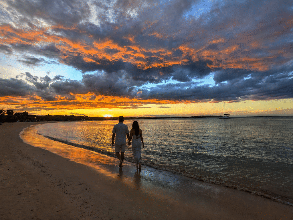 Couple walking hand in hand along Pasikuda Beach at sunset, with dramatic orange clouds, calm sea and a sailboat in the distance.